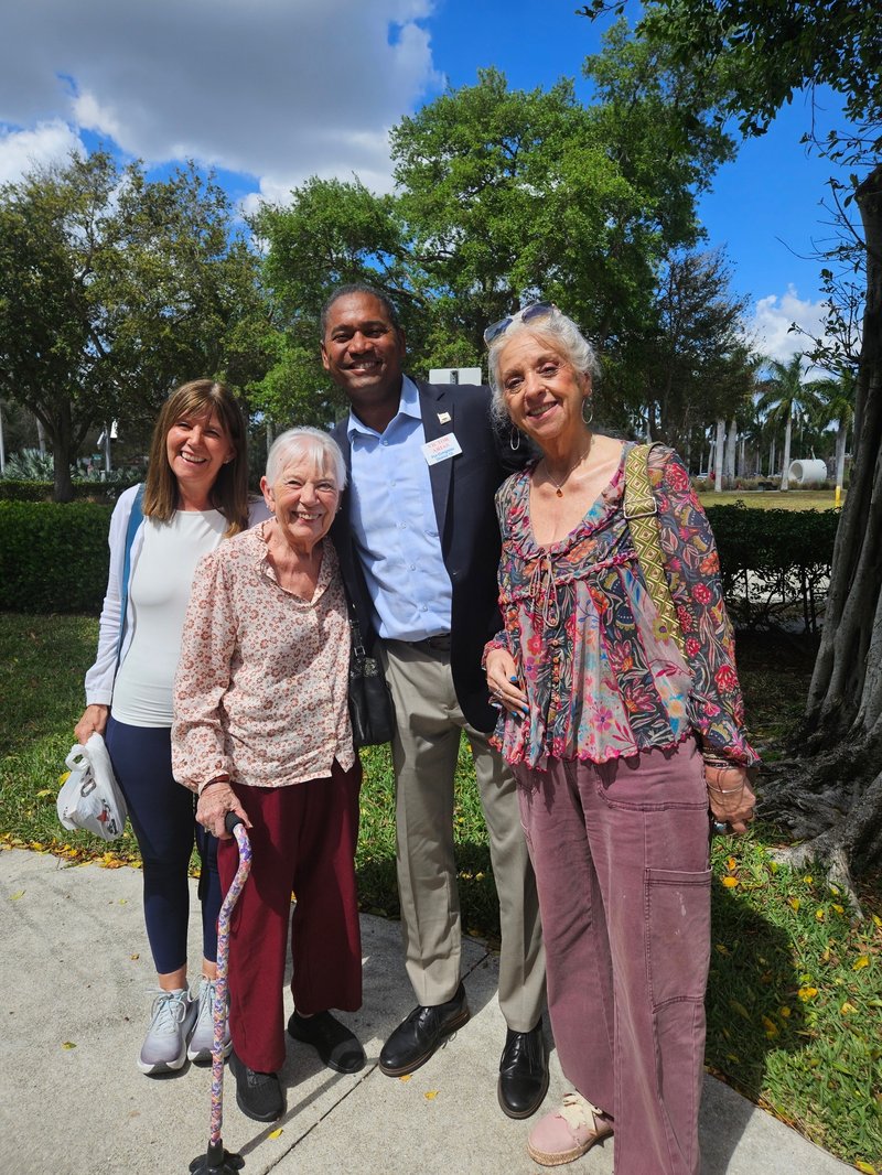 Four adults smile together outdoors beneath trees.