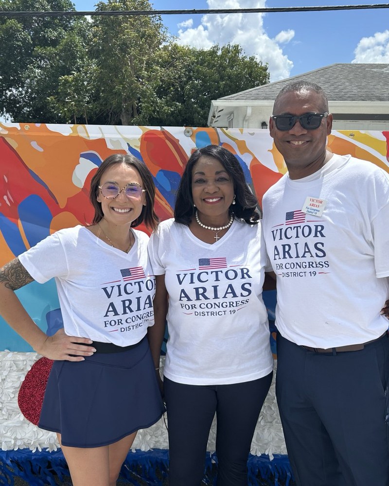 Three adults wearing Victor Arias for Congress District 19 shirts pose in front of a colorful mural.