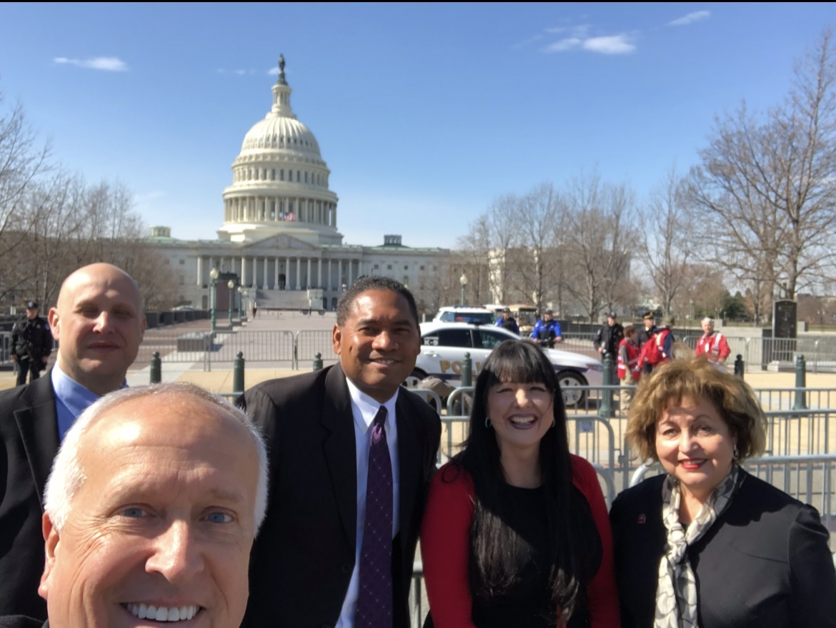 Five adults take a group photo outdoors with a domed government building behind them.
