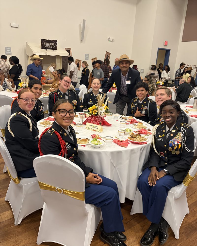 A man in a hat stands behind eight people in dark uniforms seated around a decorated banquet table.